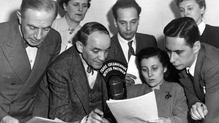“Radio Broadcast Sent To Russia By State Department” photograph from the National Archives, Harry S. Truman Library and Museum. Description: Interior view of seven men and women taken during a radio broadcast sent to Russia from the State Department’s studios in New York. Identified as left to right: Boris Brodenov, Kathrine Elene, James Shigorin, Vladmir Postman, Mrs. Lucy Bates, Victor Franzusoff, and Mrs. Tatiana Hecker, all American citizens. Lettering on top of microphone is in Russian language. (Charles Thayer supervised the programs.) Date(s): ca. February 1947.
